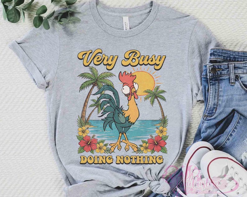 May include: A light grey t-shirt featuring a cartoon rooster design. The text "Very Busy Doing Nothing" is in yellow and orange. The graphic includes palm trees, a sun, and flowers. The shirt is styled with blue jeans and white trainers.