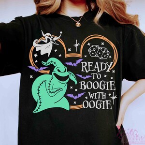 May include: A black short-sleeve t-shirt featuring a graphic of a green Oogie Boogie character, a white Zero ghost dog, dice, and purple bats. The design includes the text "READY TO BOOGIE WITH OOGIE" in white letters inside an orange Mickey ear outline.