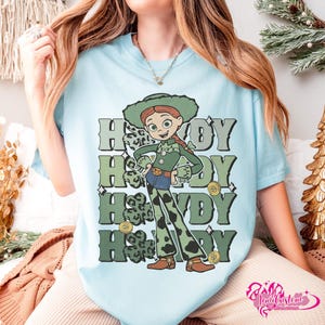 May include: Light blue t-shirt featuring a cartoon cowgirl character with the word "Howdy" repeated in green and gray. The cowgirl wears a green hat, vest, and pants with a leopard print design. Gold coins and shamrocks are also featured.