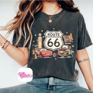 May include: Dark gray t-shirt featuring a Route 66 graphic with the text "Route 66" and the number "66". The graphic includes cartoon cars, a gas pump, and a motel sign. The shirt has a vintage, worn look.