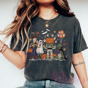K&ouml;nnte beinhalten: Dunkelgraues T-Shirt mit einem Halloween-Motiv. Das Design zeigt Zeichentrickfiguren in Kost&uuml;men, darunter Woody, Buzz Lightyear und ein Dinosaurier, mit K&uuml;rbissen, Geistern und Flederm&auml;usen. Das T-Shirt hat einen Vintage-Look.