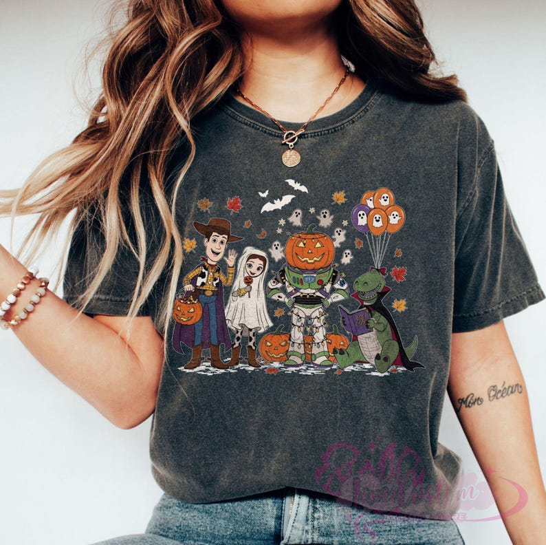 K&ouml;nnte beinhalten: Dunkelgraues T-Shirt mit einem Halloween-Motiv. Das Design zeigt Zeichentrickfiguren in Kost&uuml;men, darunter Woody, Buzz Lightyear und ein Dinosaurier, mit K&uuml;rbissen, Geistern und Flederm&auml;usen. Das T-Shirt hat einen Vintage-Look.