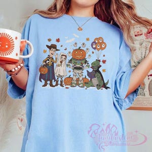 K&ouml;nnte beinhalten: Hellblaues T-Shirt mit einem Halloween-Motiv. Das Design zeigt Cartoon-Figuren in Kost&uuml;men, darunter Woody, eine Braut, ein Astronaut mit K&uuml;rbiskopf und ein lesender Dinosaurier. Orangefarbene und violette Luftballons sowie Herbstbl&auml;tter sind ebenfalls abgebildet.
