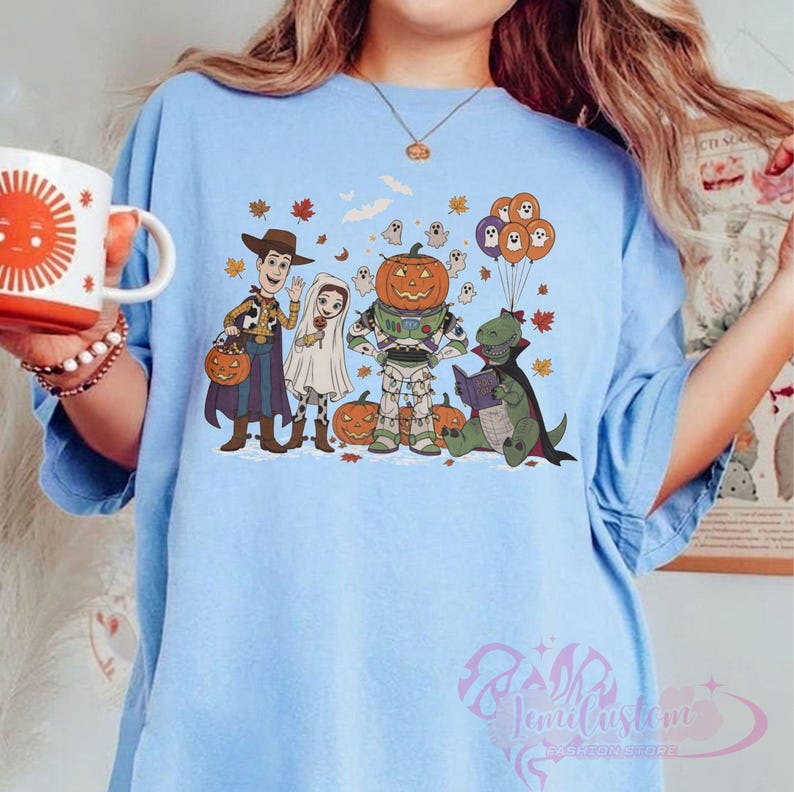 K&ouml;nnte beinhalten: Hellblaues T-Shirt mit einem Halloween-Motiv. Das Design zeigt Cartoon-Figuren in Kost&uuml;men, darunter Woody, eine Braut, ein Astronaut mit K&uuml;rbiskopf und ein lesender Dinosaurier. Orangefarbene und violette Luftballons sowie Herbstbl&auml;tter sind ebenfalls abgebildet.