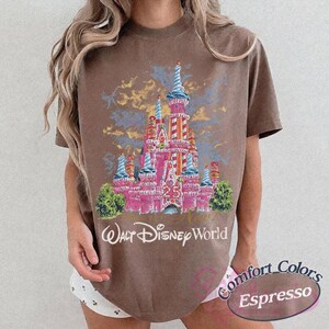 Op de afbeelding: Bruin t-shirt met een roze kasteelontwerp, blauwe en gele accenten, het nummer 25 en de woorden "Walt Disney World". Het shirt heeft ook de woorden "Comfort Colors Espresso" erop gedrukt.