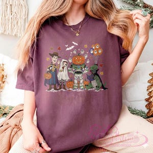 K&ouml;nnte beinhalten: Ein weinrotes T-Shirt mit einem Halloween-Motiv. Die Grafik zeigt Zeichentrickfiguren, die sich als Woody, eine Braut, ein k&uuml;rbisk&ouml;pfiger Buzz Lightyear und ein Buch lesender Dinosaurier verkleidet haben. K&uuml;rbisse, Geister und Flederm&auml;use sind ebenfalls enthalten.
