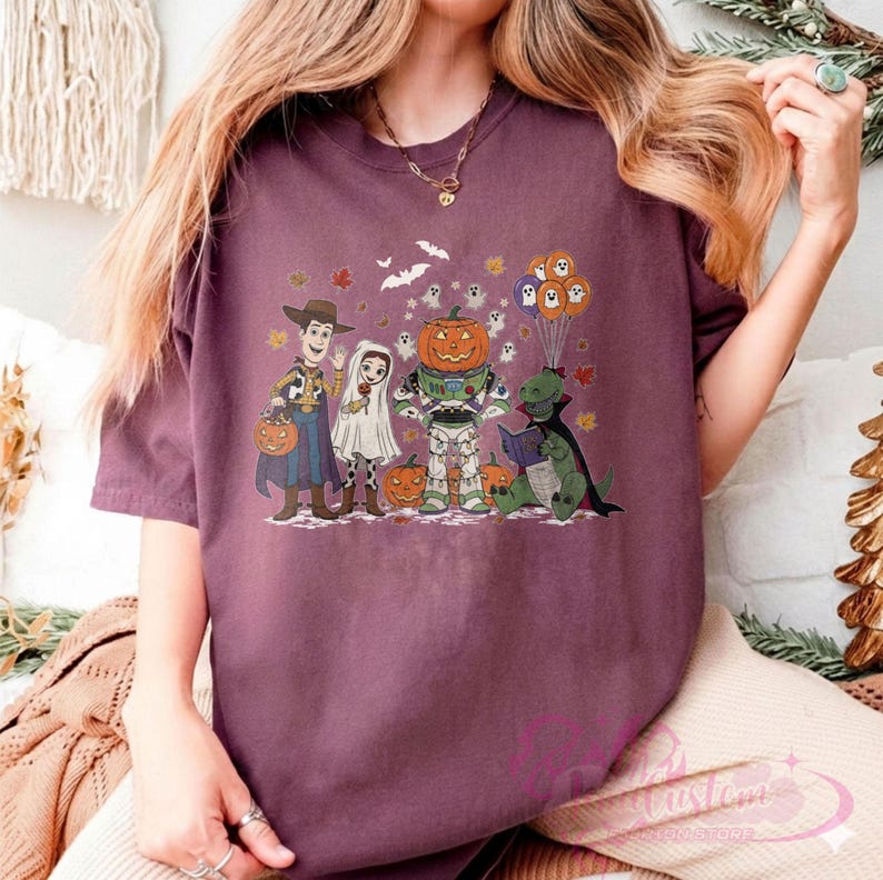 K&ouml;nnte beinhalten: Ein weinrotes T-Shirt mit einem Halloween-Motiv. Die Grafik zeigt Zeichentrickfiguren, die sich als Woody, eine Braut, ein k&uuml;rbisk&ouml;pfiger Buzz Lightyear und ein Buch lesender Dinosaurier verkleidet haben. K&uuml;rbisse, Geister und Flederm&auml;use sind ebenfalls enthalten.
