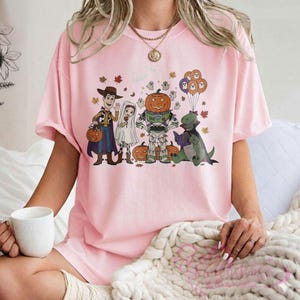 K&ouml;nnte beinhalten: Hellrosa T-Shirt mit einem Halloween-Motiv. Das Design zeigt Cartoon-Figuren in Kost&uuml;men, darunter Woody, Buzz Lightyear und ein Dinosaurier, der ein Buch liest. K&uuml;rbisse und Herbstbl&auml;tter sind ebenfalls enthalten.