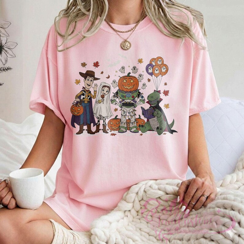 K&ouml;nnte beinhalten: Hellrosa T-Shirt mit einem Halloween-Motiv. Das Design zeigt Cartoon-Figuren in Kost&uuml;men, darunter Woody, Buzz Lightyear und ein Dinosaurier, der ein Buch liest. K&uuml;rbisse und Herbstbl&auml;tter sind ebenfalls enthalten.