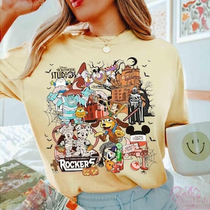 May include: A pale yellow t-shirt featuring a colorful graphic print of various animated characters in Halloween costumes, including mummies and a wizard, alongside the text Disney Hollywood Studios and Rockers.
