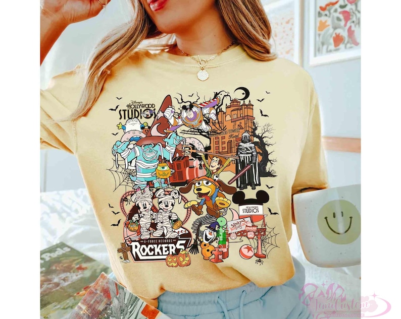 May include: A pale yellow t-shirt featuring a colorful graphic print of various animated characters in Halloween costumes, including mummies and a wizard, alongside the text Disney Hollywood Studios and Rockers.