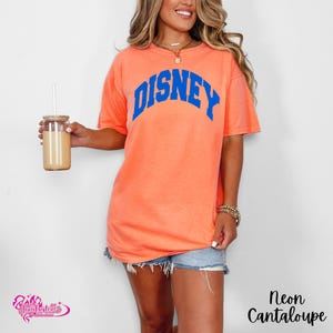 Pu&ograve; includere: T-shirt color melone neon con la scritta "DISNEY" in blu ad arco sul petto. La maglietta &egrave; di vestibilit&agrave; ampia. La modella indossa pantaloncini di jeans e una collana d'oro. Un bicchiere con una cannuccia &egrave; tenuto nella mano sinistra.