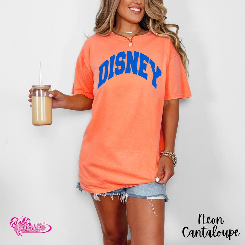 Pu&ograve; includere: T-shirt color melone neon con la scritta "DISNEY" in blu ad arco sul petto. La maglietta &egrave; di vestibilit&agrave; ampia. La modella indossa pantaloncini di jeans e una collana d'oro. Un bicchiere con una cannuccia &egrave; tenuto nella mano sinistra.