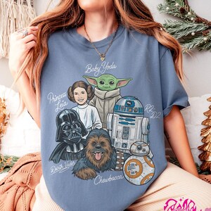 May include: A light blue t-shirt featuring a graphic of Star Wars characters, including Baby Yoda, Princess Leia, Darth Vader, Chewbacca, R2-D2, and BB-8. The names of the characters are written in a stylized font. The shirt is a comfortable, casual style.