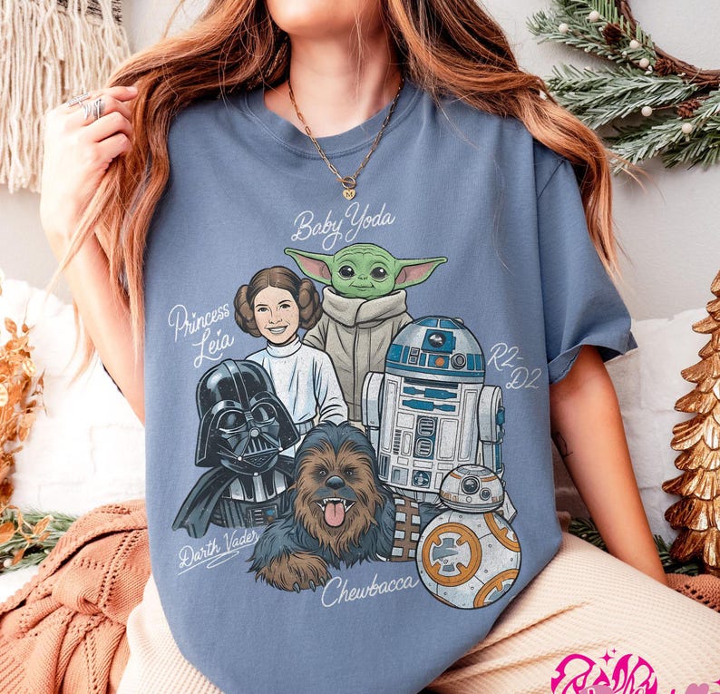 May include: A light blue t-shirt featuring a graphic of Star Wars characters, including Baby Yoda, Princess Leia, Darth Vader, Chewbacca, R2-D2, and BB-8. The names of the characters are written in a stylized font. The shirt is a comfortable, casual style.