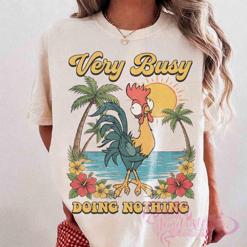 May include: Cream-coloured t-shirt with a cartoon rooster and a tropical beach scene. The shirt features the text "Very Busy Doing Nothing" in yellow and orange. Palm trees, flowers, and a sun are also part of the design.
