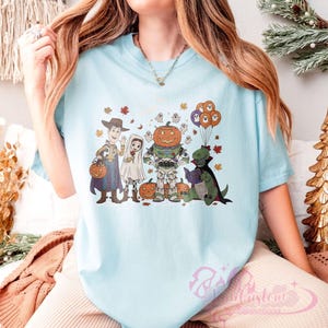 K&ouml;nnte beinhalten: Hellblaues T-Shirt mit einem Halloween-Motiv. Das Design zeigt Zeichentrickfiguren, die sich als Woody, ein Geist, ein Astronaut mit K&uuml;rbiskopf und ein Dinosaurier, der ein Buch liest, verkleidet haben. Herbstbl&auml;tter und Geisterballons vervollst&auml;ndigen den festlichen Look.
