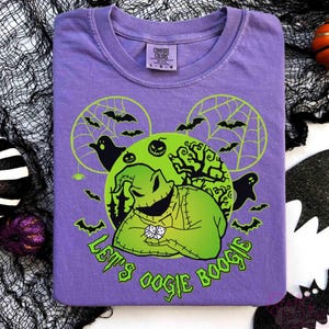May include: A purple t-shirt with a green Oogie Boogie design, bats, ghosts, and the text "Let's Oogie Boogie." The design includes a green moon and spiderwebs. Halloween decorations surround the shirt.