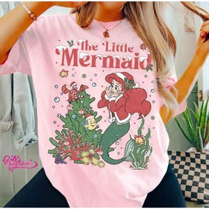 May include: Pink t-shirt featuring a Christmas-themed illustration of Ariel from The Little Mermaid. The design includes Ariel, a Christmas tree, and other characters from the movie. The text "The Little Mermaid" is displayed at the top.