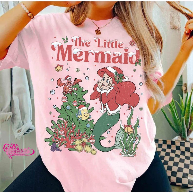 May include: Pink t-shirt featuring a Christmas-themed illustration of Ariel from The Little Mermaid. The design includes Ariel, a Christmas tree, and other characters from the movie. The text "The Little Mermaid" is displayed at the top.