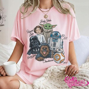 May include: Pink t-shirt featuring a graphic of Star Wars characters including Darth Vader, Princess Leia, Baby Yoda, Chewbacca, R2-D2, and BB-8. The names of the characters are written in a script font. The shirt is a light pink color.