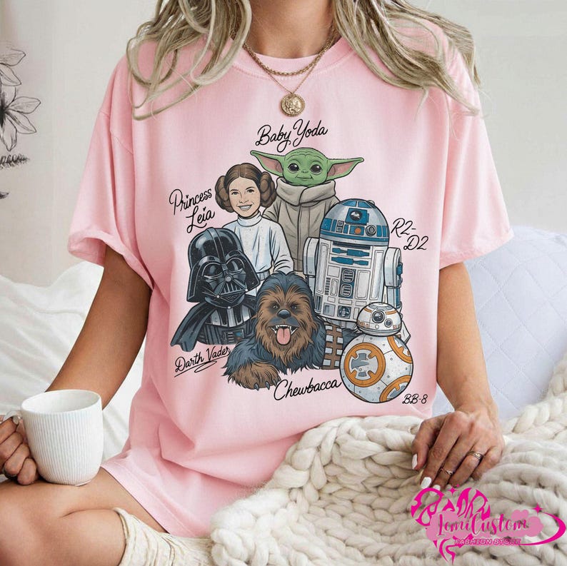May include: Pink t-shirt featuring a graphic of Star Wars characters including Darth Vader, Princess Leia, Baby Yoda, Chewbacca, R2-D2, and BB-8. The names of the characters are written in a script font. The shirt is a light pink color.