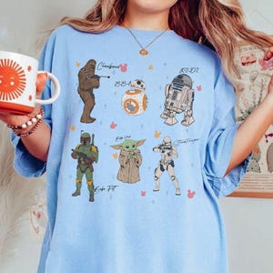 May include: Pale blue t-shirt with illustrations of Star Wars characters: Chewbacca, BB-8, R2-D2, Boba Fett, Grogu, and a Stormtrooper. The characters' names are written on the shirt.