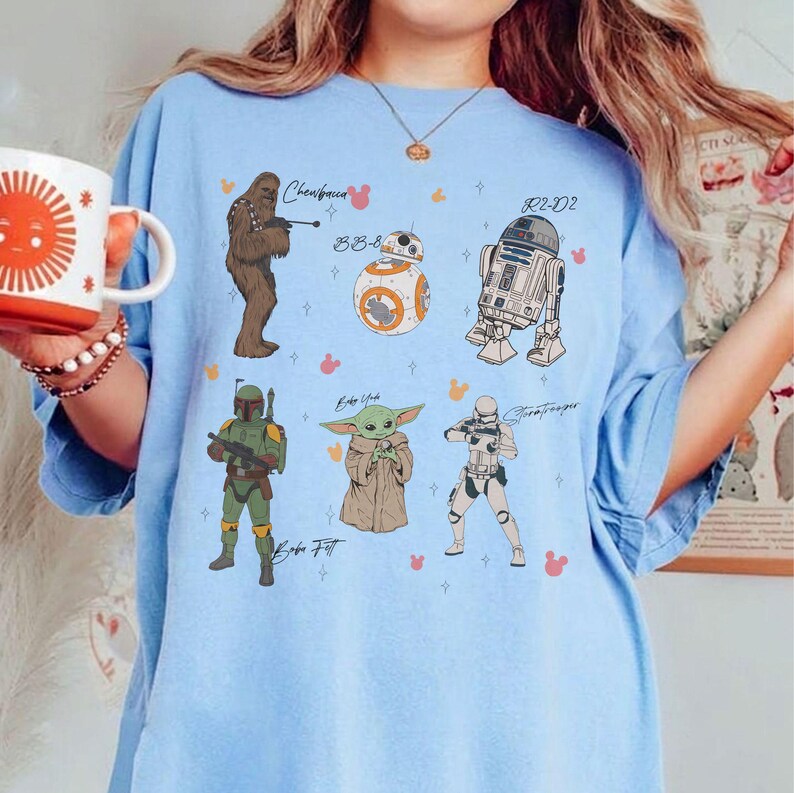 May include: Pale blue t-shirt with illustrations of Star Wars characters: Chewbacca, BB-8, R2-D2, Boba Fett, Grogu, and a Stormtrooper. The characters' names are written on the shirt.