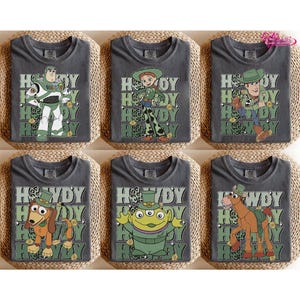 May include: Six gray t-shirts featuring characters from the Toy Story franchise. Each shirt has a different character with the word "Howdy" in a green and white design. Characters include Buzz Lightyear, Jessie, Woody, Slinky Dog, an alien, and Bullseye.