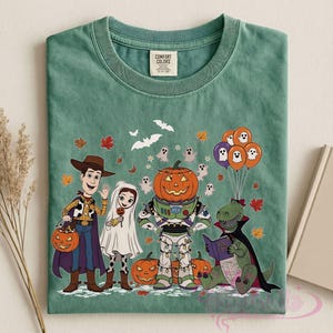 K&ouml;nnte beinhalten: Gr&uuml;nes T-Shirt mit einem Halloween-Motiv mit Cartoon-Charakteren in Kost&uuml;men. Das Design umfasst einen Cowboy, einen Geist, einen Astronauten mit K&uuml;rbiskopf und einen Dinosaurier, der ein Buch liest. K&uuml;rbisse, Geister, Flederm&auml;use und Herbstbl&auml;tter.