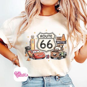 May include: Cream-colored t-shirt featuring a Route 66 graphic with the text "Route 66" and the number "66". The graphic includes cartoon cars, a gas pump, and the words "Cozy Cone Motel".