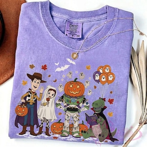 K&ouml;nnte beinhalten: Ein lavendelfarbenes Comfort Colors T-Shirt mit einem Halloween-Motiv. Das Design zeigt Cartoon-Figuren in Kost&uuml;men, K&uuml;rbisse, Geister und Herbstbl&auml;tter. Eine goldene Halskette liegt auf dem T-Shirt.