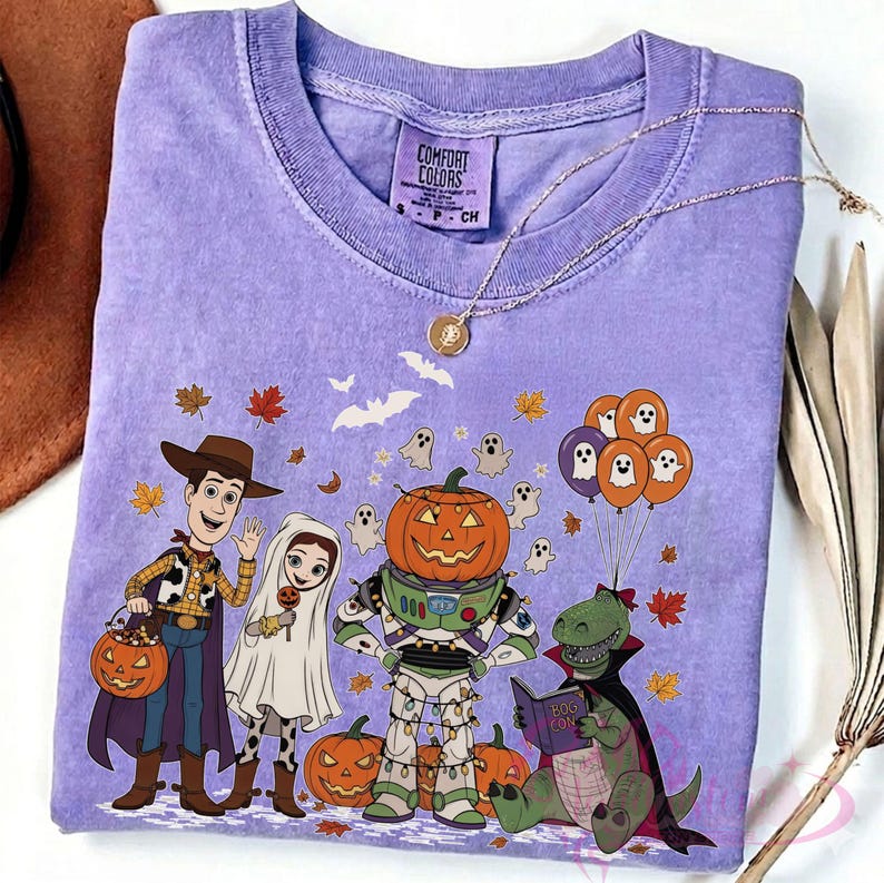 K&ouml;nnte beinhalten: Ein lavendelfarbenes Comfort Colors T-Shirt mit einem Halloween-Motiv. Das Design zeigt Cartoon-Figuren in Kost&uuml;men, K&uuml;rbisse, Geister und Herbstbl&auml;tter. Eine goldene Halskette liegt auf dem T-Shirt.