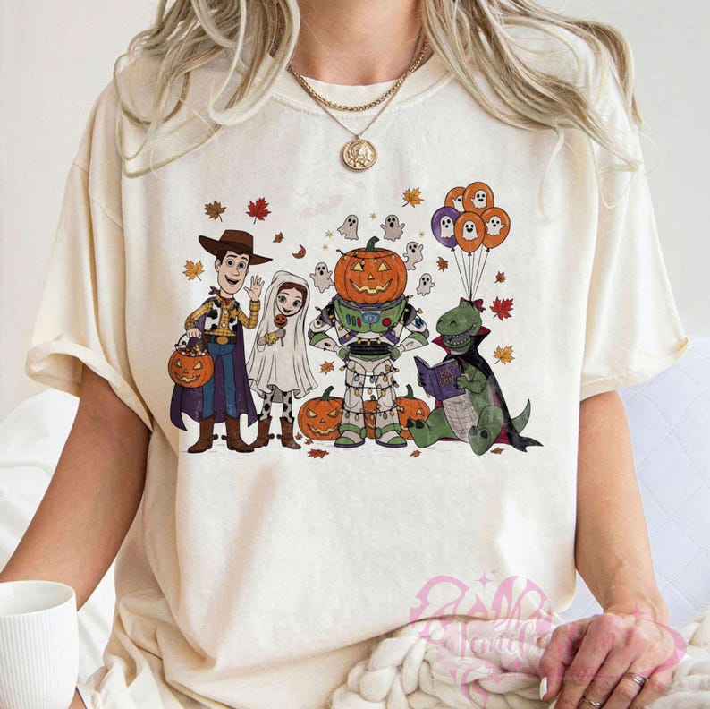 K&ouml;nnte beinhalten: Cremefarbenes T-Shirt mit einem Halloween-Motiv. Das Design zeigt Cartoon-Charaktere in Kost&uuml;men, darunter Woody, Buzz Lightyear und ein Dinosaurier, der ein Buch liest. K&uuml;rbisse, Geister und Herbstbl&auml;tter sind ebenfalls enthalten.