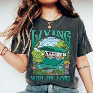 May include: Dark gray t-shirt featuring a green graphic with the text "LIVING WITH THE LAND." The graphic depicts a boat with Disney characters, surrounded by green foliage and flowers. The shirt has a vintage, worn look.