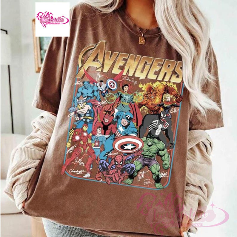 May include: Brown t-shirt featuring the word "Avengers" in gold lettering above a colorful graphic of various Marvel superheroes. The graphic includes characters like Iron Man, Captain America, and Spider-Man. The shirt has a vintage, worn look.