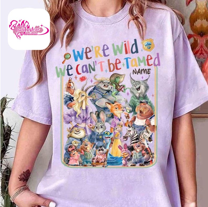 Puede incluir: Camiseta lila claro con un gr&aacute;fico colorido de varios animales de dibujos animados. El texto "We're Wild We Can't Be Tamed" se muestra encima de los personajes, con espacio para un nombre. La camiseta tiene cuello redondo y mangas cortas.