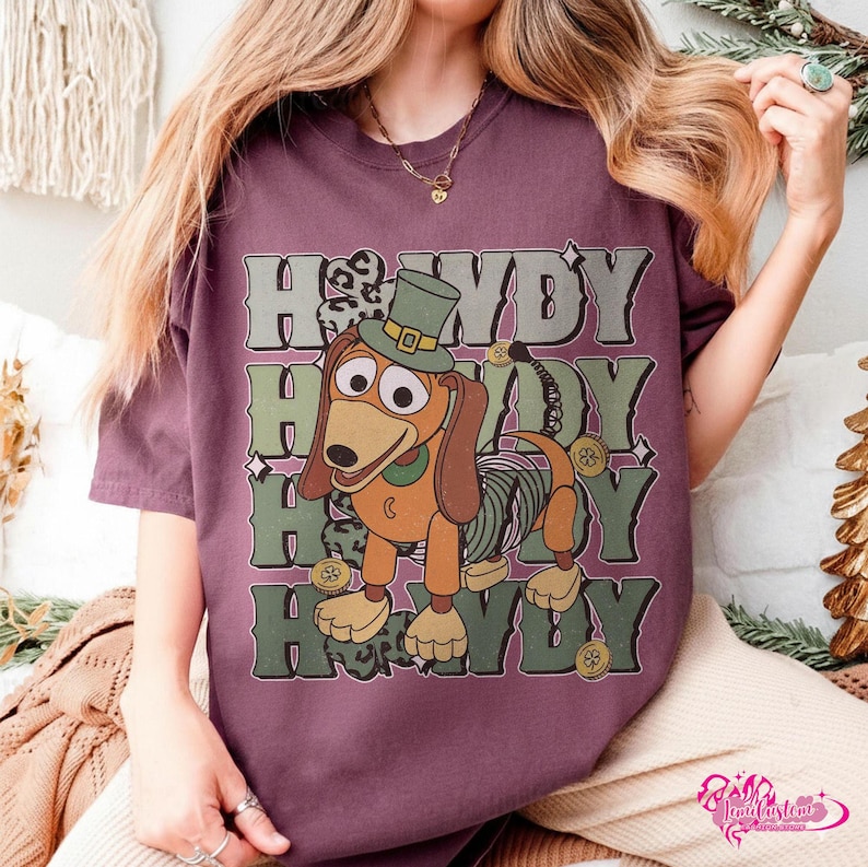 May include: A maroon t-shirt featuring a cartoon dachshund wearing a green top hat and a green and gray striped midsection. The word "Howdy" is stacked four times in green and gray block letters.