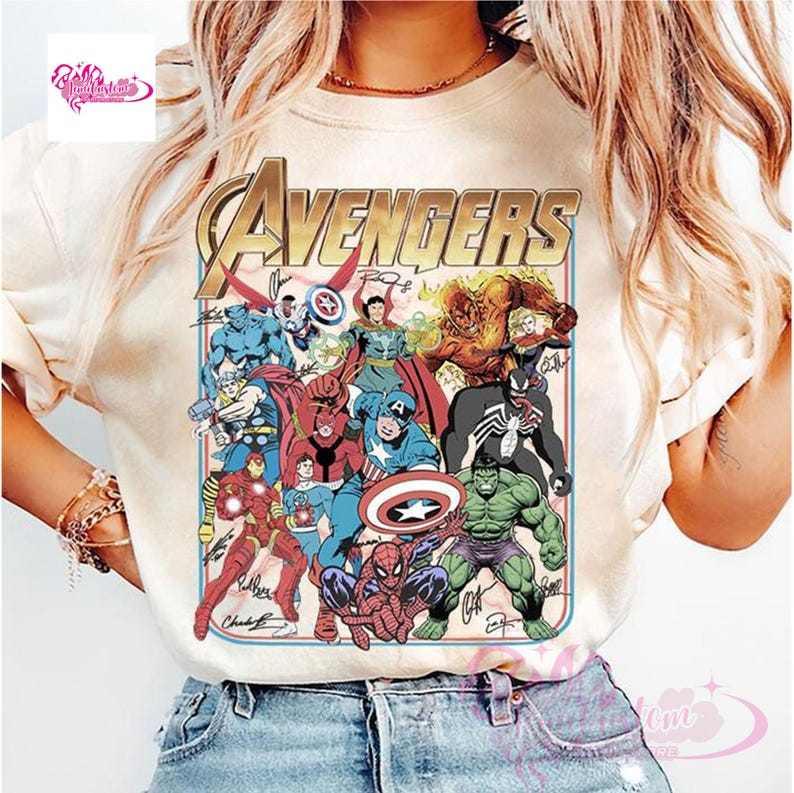 May include: Cream-colored t-shirt featuring a colorful graphic of the Avengers team. The design includes the word "AVENGERS" in gold lettering, along with various superhero characters and their signatures. The shirt is made of a soft material.