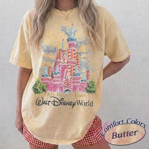 Op de afbeelding: Boterkleurig T-shirt met een kleurrijke illustratie van een kasteel met de tekst "This is Now! Walt Disney World". Het shirt heeft korte mouwen en een relaxte pasvorm. De tekst "Comfort Colors Butter" staat er ook op.