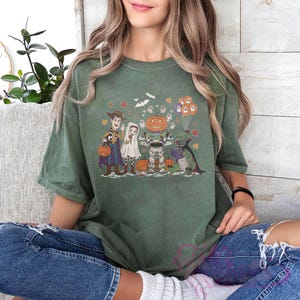 K&ouml;nnte beinhalten: Gr&uuml;nes T-Shirt mit einem Halloween-Motiv. Das Design zeigt Zeichentrickfiguren in Kost&uuml;men, darunter Woody, ein Geist, ein K&uuml;rbis und ein Dinosaurier. Die Grafik enth&auml;lt auch Flederm&auml;use, Bl&auml;tter und Geisterballons.