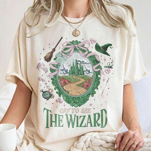 May include: Cream-colored t-shirt featuring a whimsical illustration of a green castle, a yellow brick road, and the text "OFF TO SEE THE WIZARD" in green. The design includes a witch's hat, a broom, and other decorative elements.
