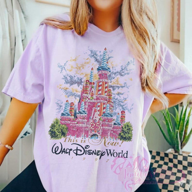 Op de afbeelding: Lichtpaarse t-shirt met een kleurrijke illustratie van een kasteel en de tekst "This is Now! Walt Disney World 1997". Het kasteel is roze met blauwe en witte details. Het t-shirt heeft korte mouwen.