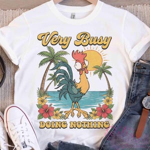 May include: White t-shirt featuring a cartoon rooster design. The rooster is yellow, blue, and red, with the text "Very Busy Doing Nothing" in yellow and black. Palm trees, a sun, and flowers surround the rooster.