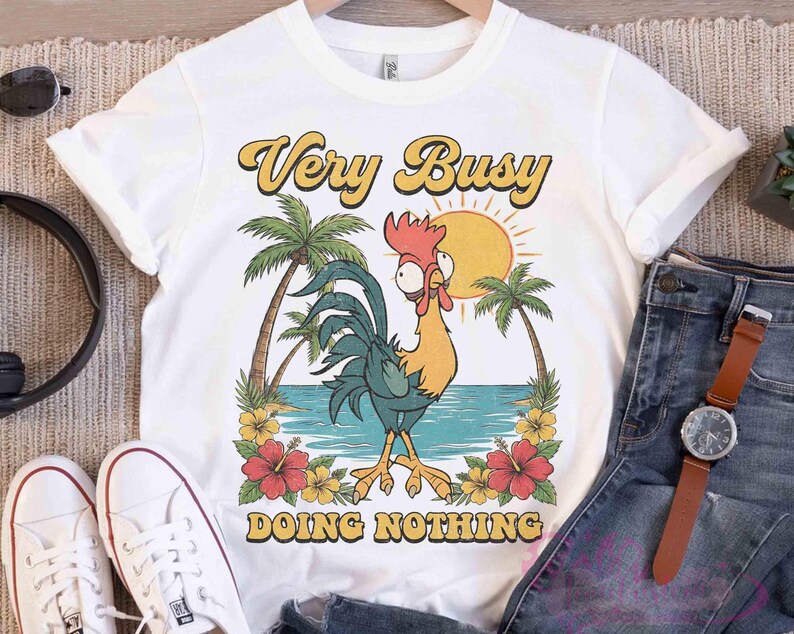 May include: White t-shirt featuring a cartoon rooster design. The rooster is yellow, blue, and red, with the text "Very Busy Doing Nothing" in yellow and black. Palm trees, a sun, and flowers surround the rooster.