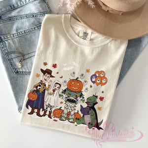 K&ouml;nnte beinhalten: Cremefarbenes T-Shirt mit einem Halloween-Motiv mit Comicfiguren in Kost&uuml;men. Das Design umfasst Woody, einen Geist, Buzz Lightyear, einen K&uuml;rbis und einen Dinosaurier. Das T-Shirt ist gefaltet und auf einer blauen Jeans platziert.