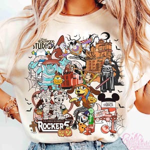 May include: A cream-colored t-shirt featuring a graphic print of various cartoon characters in Halloween costumes, including mummies, a space ranger, and a cowboy. The design includes the text Disney's Hollywood Studios, Rocker 5, and Slinky Dog.