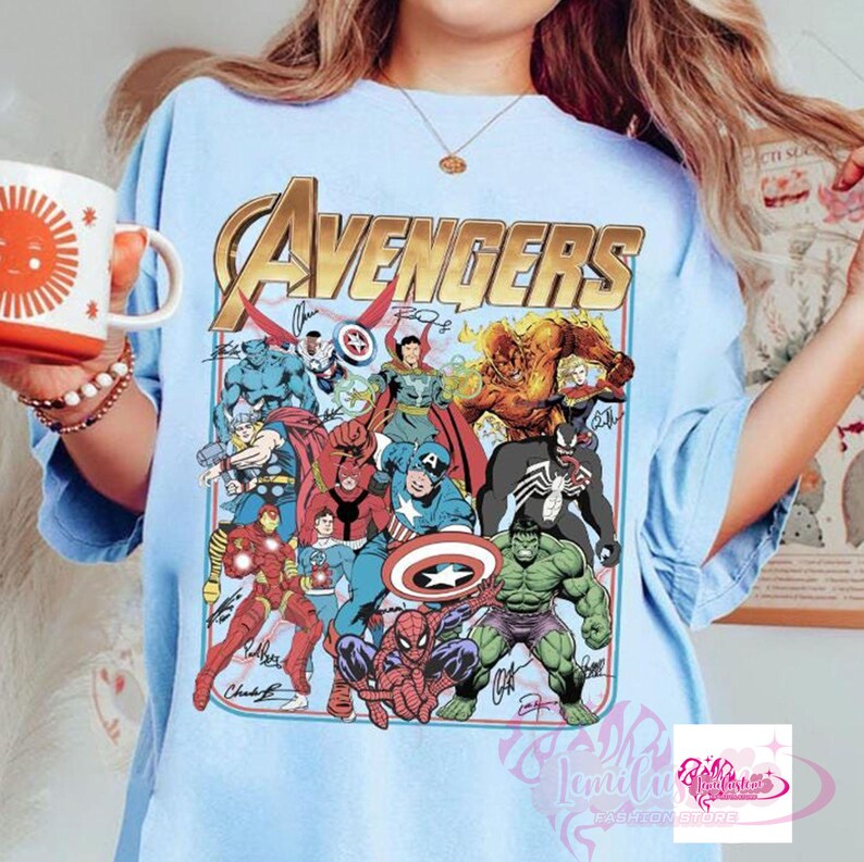 May include: Light blue t-shirt featuring the word "Avengers" in gold lettering, with a colorful graphic of various Marvel superheroes. The design includes characters like Iron Man, Spider-Man, and Captain America. The shirt has a crew neck and short sleeves.