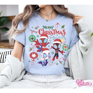 May include: Light blue t-shirt featuring a festive "Merry Christmas" design. The graphic includes Spider-Man characters wearing Santa hats and Christmas ornaments. The design is surrounded by snowflakes and holly.