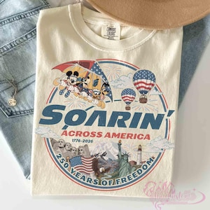 May include: Cream-colored t-shirt featuring a patriotic design with Mickey Mouse, Donald Duck, and Goofy. The graphic includes hot air balloons, fireworks, and the text "Soarin' Across America" with the years 1776-2026 and "250 Years of Freedom".